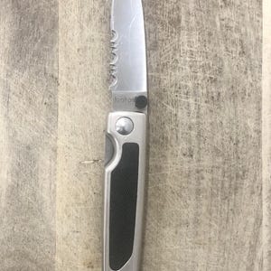 Pode incluir: Uma faca dobr&aacute;vel prateada com uma l&acirc;mina parcialmente serrilhada. A pega &eacute; prateada com uma pega de borracha preta. A palavra "Kershaw" est&aacute; impressa na l&acirc;mina. A faca &eacute; mostrada numa superf&iacute;cie de madeira.