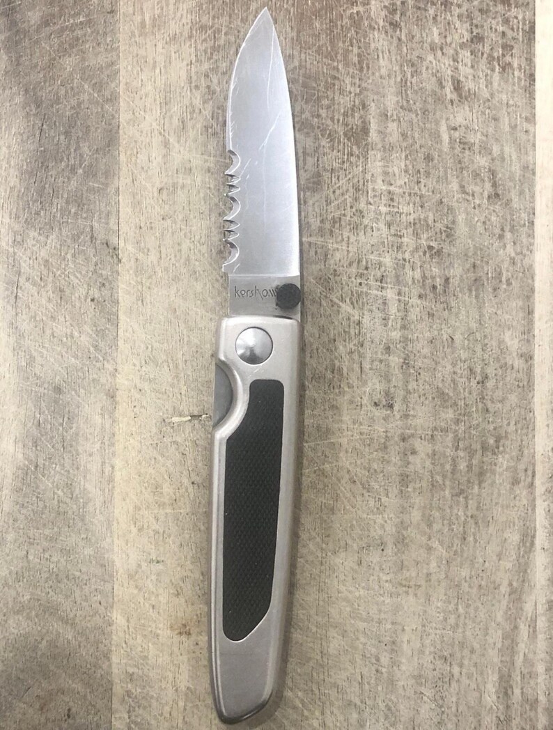 Pode incluir: Uma faca dobr&aacute;vel prateada com uma l&acirc;mina parcialmente serrilhada. A pega &eacute; prateada com uma pega de borracha preta. A palavra "Kershaw" est&aacute; impressa na l&acirc;mina. A faca &eacute; mostrada numa superf&iacute;cie de madeira.