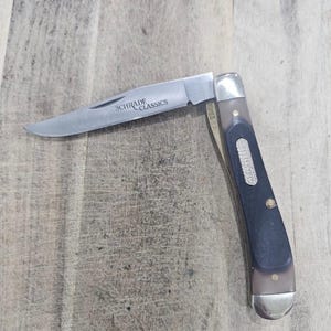 May include: A Schrade Classics folding knife with a partially open, silver blade. The handle is black and brown with a silver end cap. The knife is resting on a light brown, textured surface.