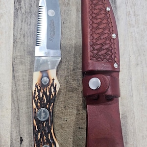 Puede incluir: Una navaja plegable Remington R-6 de acero inoxidable con borde dentado y mango de hueso. La navaja está junto a una funda de cuero marrón con un patrón decorativo y cierre de botón a presión. Sobre una superficie de madera.