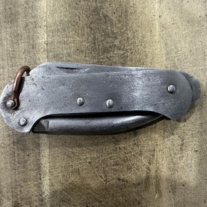 Pode incluir: Um canivete de bolso fechado com um cabo de metal cinzento desgastado, fixado por quatro rebites. Um pequeno anel de metal cor de cobre está preso a uma extremidade do cabo. A superfície do metal apresenta um acabamento texturado e usado.