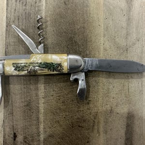 May include: A multi-tool pocket knife with a cream-colored handle featuring a green landscape illustration and the text Katoomba Blue Mountains. The metal tool includes a main blade, a corkscrew, a bottle opener, and a small flat-head driver.