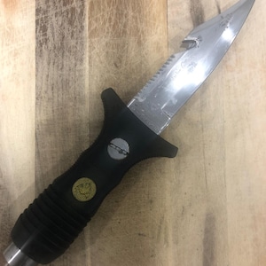 May include: A stainless steel dive knife with a serrated edge and a black rubber handle. The handle features a gold emblem and a silver end cap. The blade has the text "Sea Lion" etched on it.