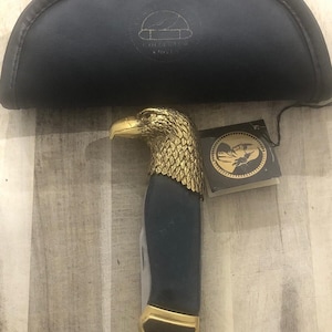 Könnte beinhalten: Ein dekoratives Taschenmesser mit einem Adlerkopfgriff. Der Griff ist gold und blau, mit einer goldenen Klinge. Das Messer befindet sich in einem schwarzen Etui mit der Aufschrift "COLLECTOR KNIVES". Ein Etikett mit einem Fischlogo ist angebracht.