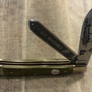 Op de afbeelding: Een vouwmes met een groene handgreep en gouden details. Het gedeeltelijk geopende lemmet toont een ge&euml;tst ontwerp en de tekst "U.S. MARINES". Het mes heeft een klein embleem op het handvat.