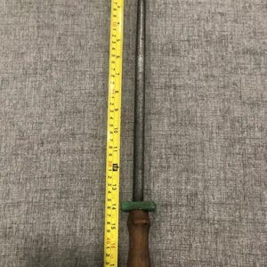 May include: A steel knife sharpening rod with a wooden handle. The rod is approximately 15 inches long, with a green guard and a loop at the end of the handle. A yellow measuring tape is next to the rod.