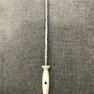 Puede incluir: Un afilador de cuchillos de acero plateado con un mango blanco y un anillo negro en la base. El afilador está diseñado para afilar el filo de los cuchillos. El mango tiene un agujero para colgar.