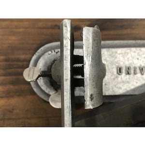 May include: Close-up of a vintage metal clamp with a screw mechanism. The clamp is silver-toned with visible wear and tear, suggesting age. The word "UNIV" is partially visible on the background. The clamp is likely used for woodworking or similar tasks.