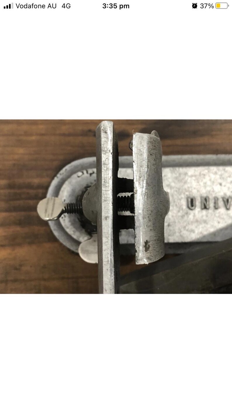 May include: Close-up of a vintage metal clamp with a screw mechanism. The clamp is silver-toned with visible wear and tear, suggesting age. The word "UNIV" is partially visible on the background. The clamp is likely used for woodworking or similar tasks.