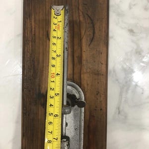 May include: A vintage metal and wood tool with a yellow measuring tape. The tape shows measurements in inches, with the tool measuring approximately 9 inches in length. The wooden base is a dark brown color.
