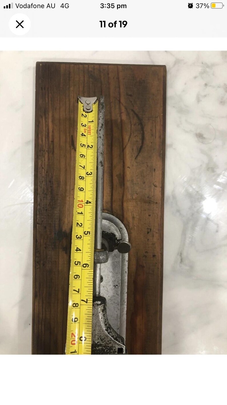 May include: A vintage metal and wood tool with a yellow measuring tape. The tape shows measurements in inches, with the tool measuring approximately 9 inches in length. The wooden base is a dark brown color.