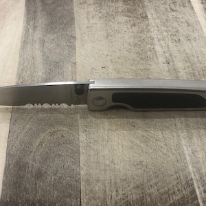 Pode incluir: Uma faca dobr&aacute;vel de a&ccedil;o inoxid&aacute;vel com uma l&acirc;mina parcialmente serrilhada. A pega &eacute; prateada com uma pega de borracha preta. A faca &eacute; mostrada aberta, mostrando a aresta da l&acirc;mina e o design geral.
