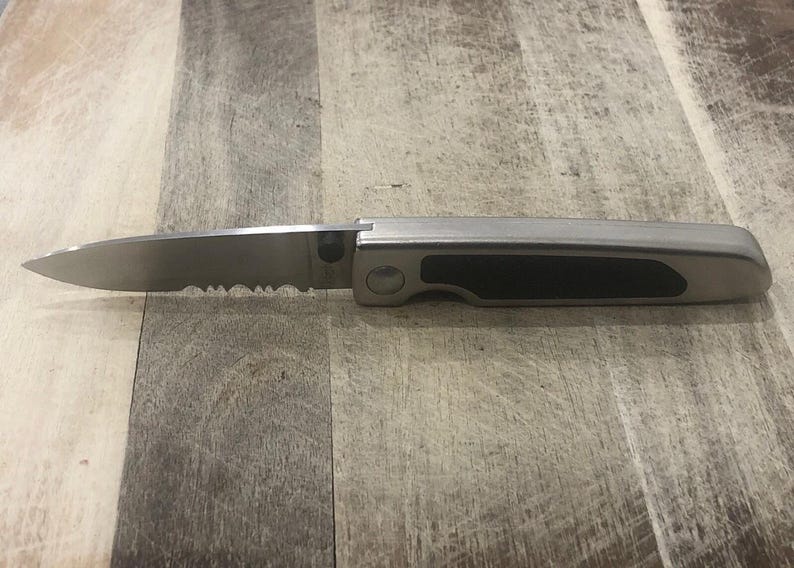Pode incluir: Uma faca dobr&aacute;vel de a&ccedil;o inoxid&aacute;vel com uma l&acirc;mina parcialmente serrilhada. A pega &eacute; prateada com uma pega de borracha preta. A faca &eacute; mostrada aberta, mostrando a aresta da l&acirc;mina e o design geral.