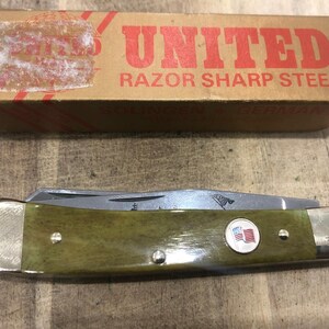 Op de afbeelding: Een vintage zakmes met een groene handgreep en een zilveren lemmet. De handgreep heeft een cirkelvormig embleem met een Amerikaanse vlag. Het mes ligt naast een kartonnen doos met het woord "UNITED" in rood.