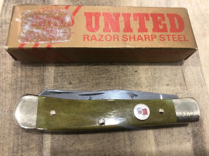 Op de afbeelding: Een vintage zakmes met een groene handgreep en een zilveren lemmet. De handgreep heeft een cirkelvormig embleem met een Amerikaanse vlag. Het mes ligt naast een kartonnen doos met het woord "UNITED" in rood.