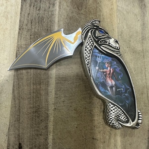 May include: A folding pocket knife with a silver-toned handle featuring a sculpted dragon head with blue eyes and an inset illustration of a figure holding a trident. The blade is shaped like a bat wing with gold-coloured accents on the metal surface.