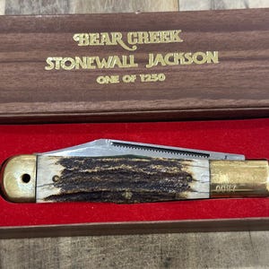 Könnte beinhalten: Ein Klappmesser mit strukturiertem Hirschhorngriff und Messingbacken, das in einer braunen Schachtel mit rotem Samtfutter liegt. Der Deckel der Schachtel trägt den goldenen Schriftzug BEAR CREEK STONEWALL JACKSON ONE OF 1250.