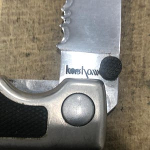 May include: Close-up of a partially open folding knife with a serrated blade. The blade is silver with the brand name "Kershaw" printed on it. The handle is black and silver.