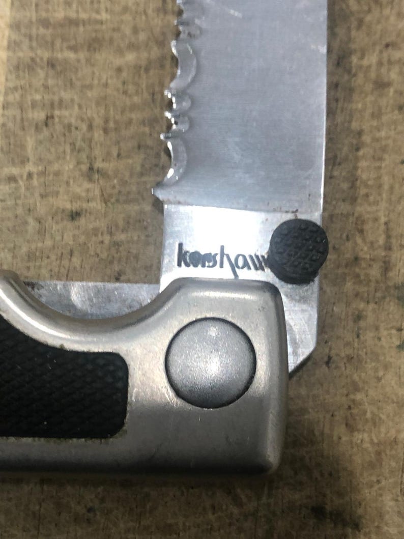 May include: Close-up of a partially open folding knife with a serrated blade. The blade is silver with the brand name "Kershaw" printed on it. The handle is black and silver.