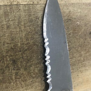May include: Close-up of a serrated stainless steel knife blade. The blade has a sharp point and a partially serrated edge. The word "Kershaw" is visible on the blade. The knife is set against a wooden surface.