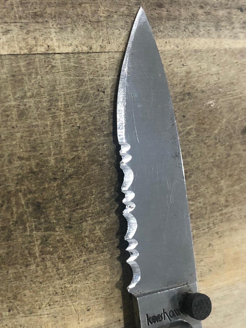 May include: Close-up of a serrated stainless steel knife blade. The blade has a sharp point and a partially serrated edge. The word "Kershaw" is visible on the blade. The knife is set against a wooden surface.