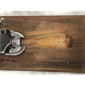 May include: Vintage can opener with a wooden handle and a black and silver metal mechanism, mounted on a dark wooden board. The opener shows signs of wear, suggesting it's an antique kitchen tool.