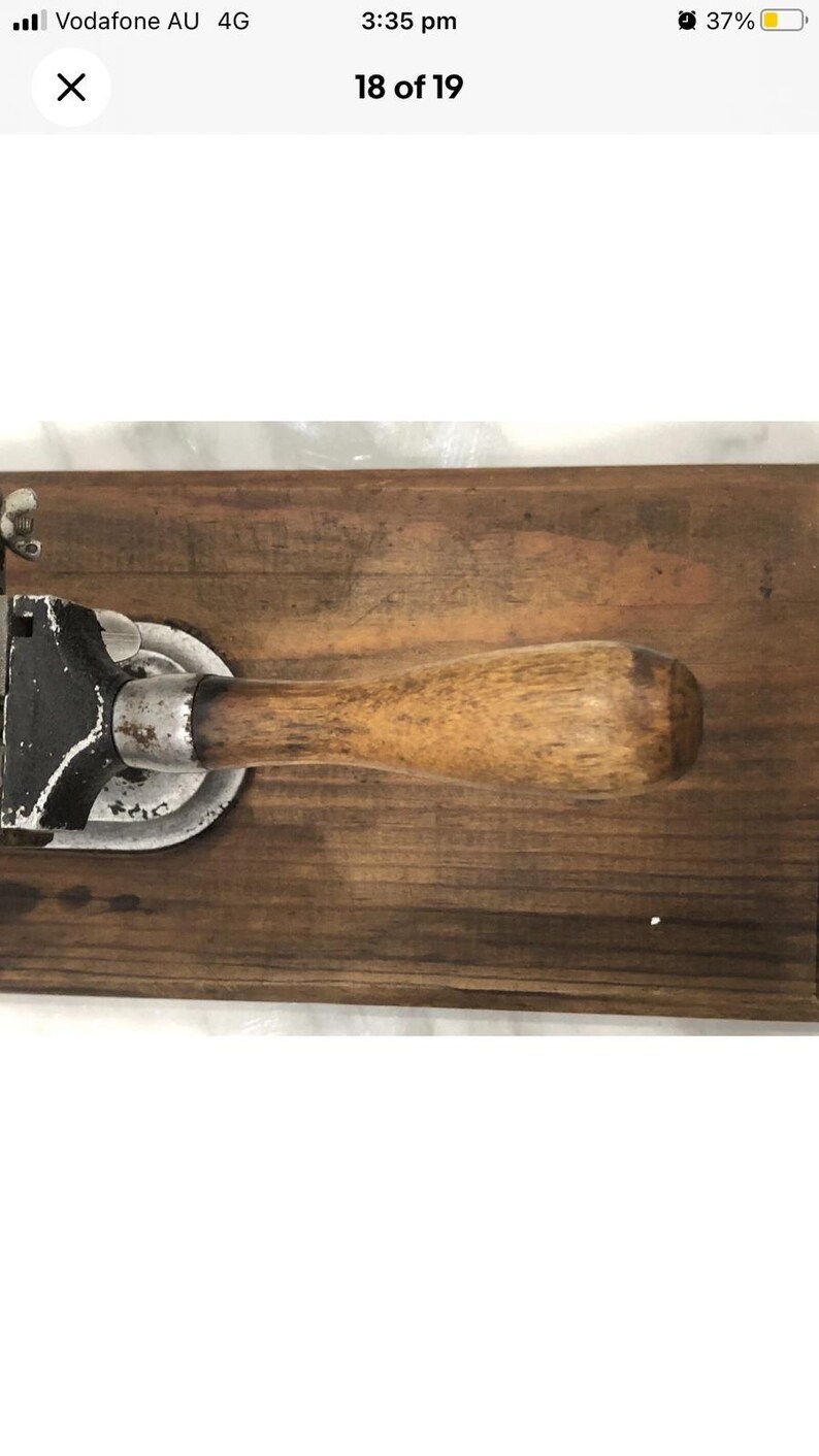 May include: Vintage can opener with a wooden handle and a black and silver metal mechanism, mounted on a dark wooden board. The opener shows signs of wear, suggesting it's an antique kitchen tool.