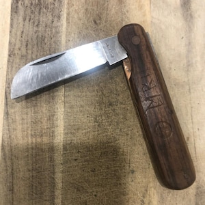 May include: A partially open pocket knife with a wooden handle and a silver blade. The blade is marked with the text "SHREBHAM". The handle is engraved with the initials "MR" and a circular symbol. The knife rests on a wooden surface.