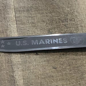 Op de afbeelding: Een zilverkleurig mesblad met de tekst "U.S. MARINES" gegraveerd. Het blad heeft ook drie sterren en het Marine Corps-embleem. Het mes ligt op een houten achtergrond.