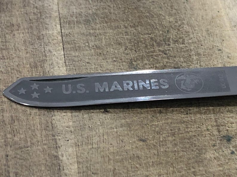 Op de afbeelding: Een zilverkleurig mesblad met de tekst "U.S. MARINES" gegraveerd. Het blad heeft ook drie sterren en het Marine Corps-embleem. Het mes ligt op een houten achtergrond.