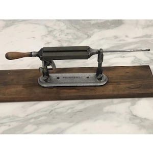 May include: An antique knife sharpener with a wooden handle and a metal frame. The sharpener is mounted on a wooden base and has a long, thin metal rod extending from one end. The metal frame is dark gray.