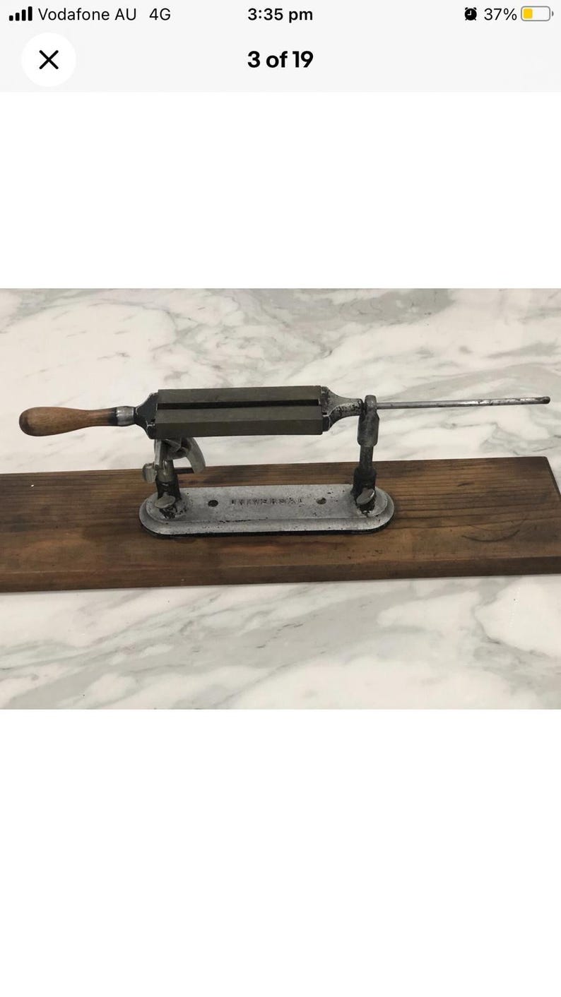 May include: An antique knife sharpener with a wooden handle and a metal frame. The sharpener is mounted on a wooden base and has a long, thin metal rod extending from one end. The metal frame is dark gray.