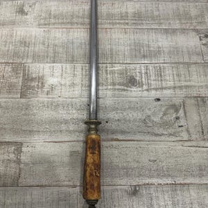May include: A sharpening steel featuring a long, tapered, metallic rod with a textured, light brown handle. The handle has a weathered, organic appearance and is capped with a small, dark metal ring at the base for hanging or storage purposes.