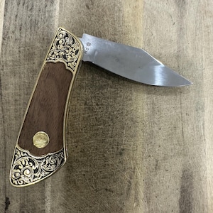 Puede incluir: Una navaja plegable con una hoja de acero de tono plateado marcada con las letras FM. El mango presenta incrustaciones de madera marrón oscura, virolas de metal dorado con grabados de volutas negras y un pequeño emblema dorado circular.