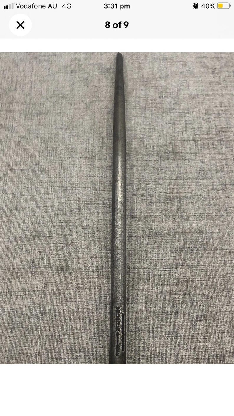 May include: A long, slender, metal tool with a pointed end. The tool is dark gray and appears to be made of steel. The tool has a textured surface and a small inscription near the base.