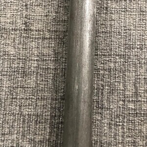 May include: A close-up of a cylindrical metal rod with a slightly textured surface. The rod is a dull gray color and appears to be made of steel or a similar metal. The background is a gray, textured fabric.