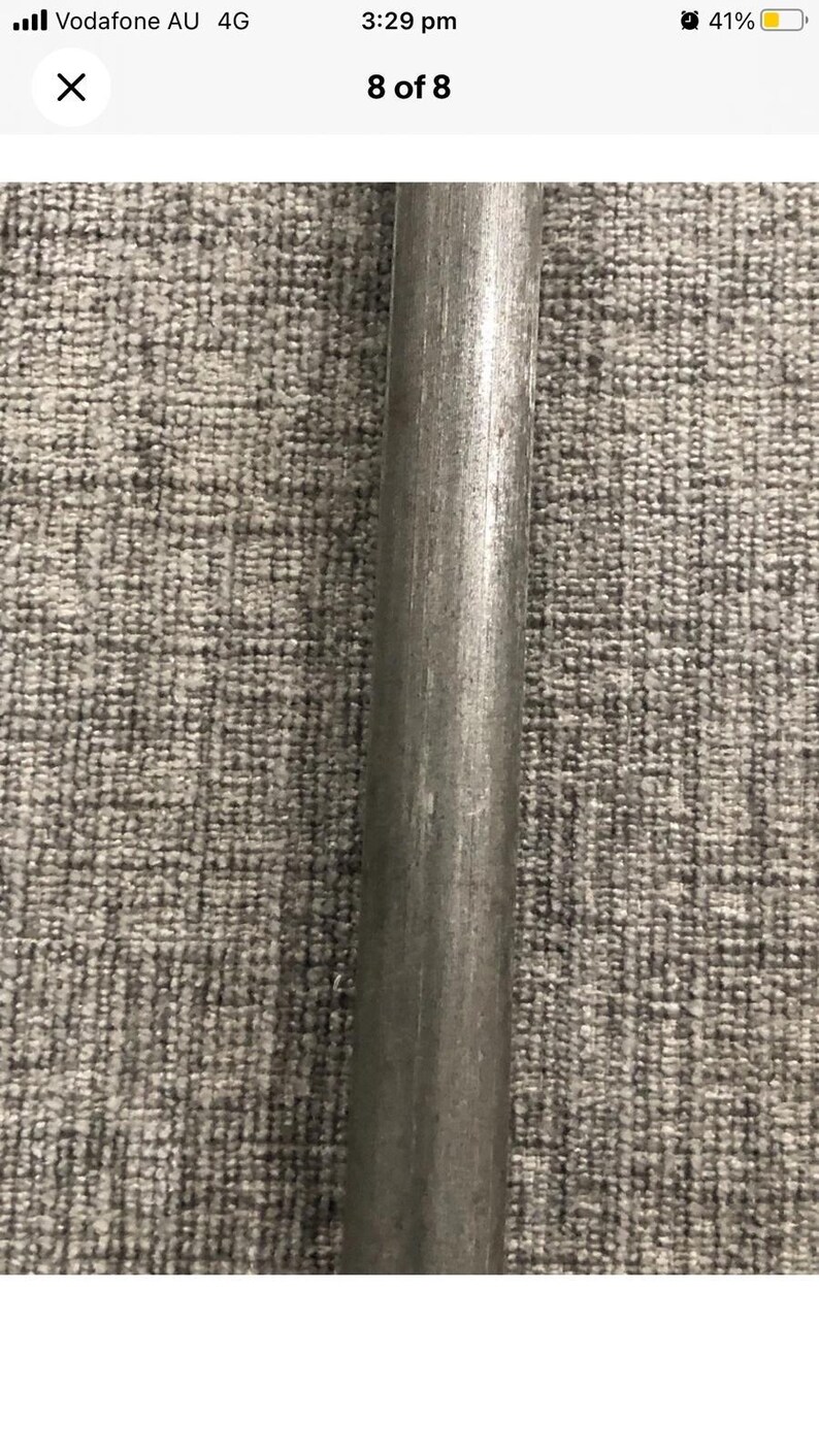 May include: A close-up of a cylindrical metal rod with a slightly textured surface. The rod is a dull gray color and appears to be made of steel or a similar metal. The background is a gray, textured fabric.