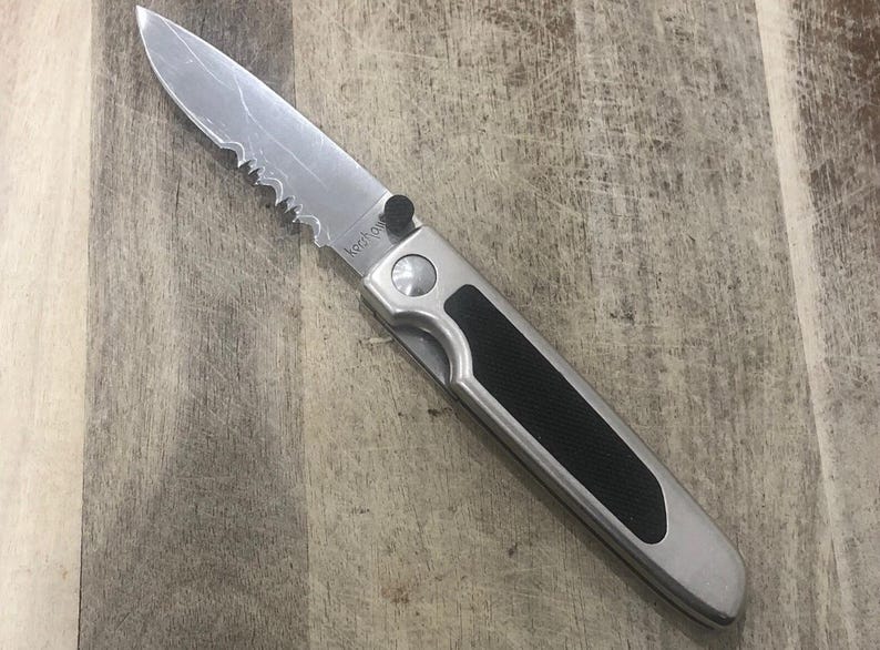 Pode incluir: Uma faca dobr&aacute;vel prateada com uma l&acirc;mina parcialmente serrilhada e uma pega preta. A faca est&aacute; sobre uma superf&iacute;cie de madeira. A marca "Kershaw" &eacute; vis&iacute;vel na l&acirc;mina.