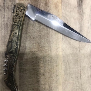 May include: A folding pocket knife with a silver blade and a decorative handle. The handle features an embossed fish design and a built-in corkscrew. The blade is marked "Pakistan".