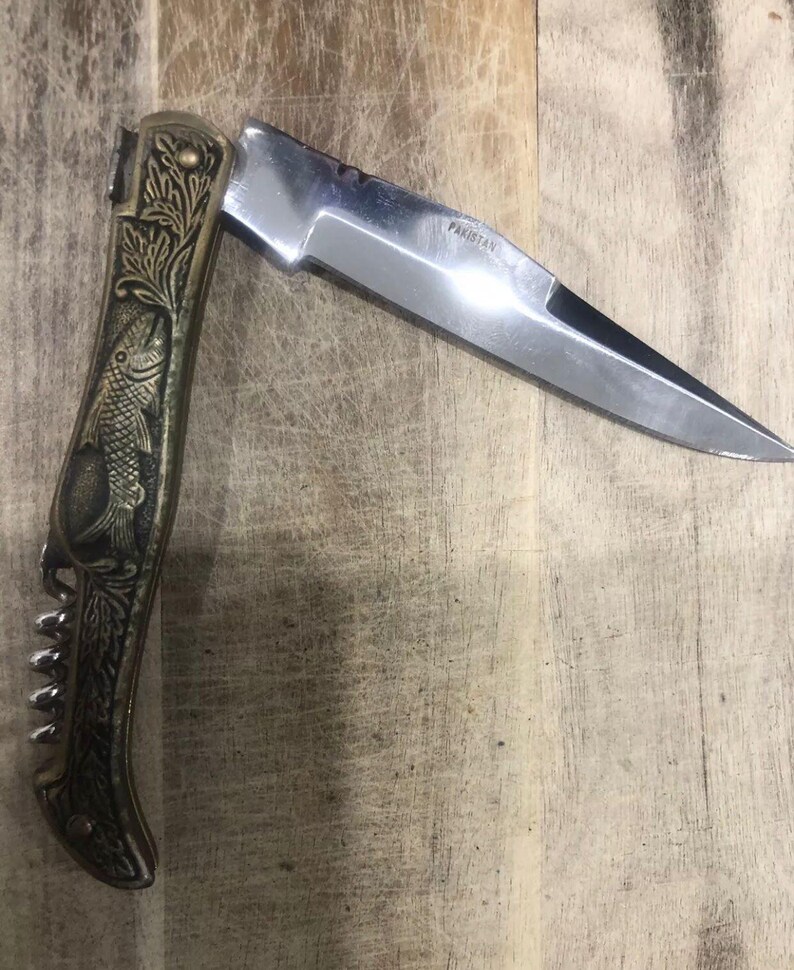 May include: A folding pocket knife with a silver blade and a decorative handle. The handle features an embossed fish design and a built-in corkscrew. The blade is marked "Pakistan".