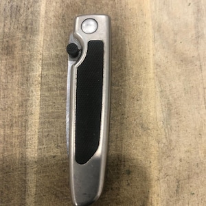 May include: A silver folding knife with a black textured grip. The knife has a button on the side and a circular button on the top. The blade is folded inside the handle.