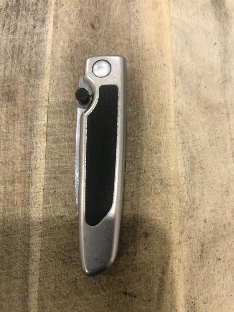 May include: A silver folding knife with a black textured grip. The knife has a button on the side and a circular button on the top. The blade is folded inside the handle.