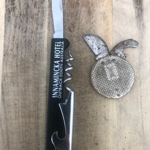 May include: A black waiter's corkscrew with a silver blade, corkscrew, and bottle opener, featuring the white text INNAMINCKA HOTEL OUTBACK SOUTH AUSTRALIA, alongside a round, textured silver-toned metal pocket knife with two small blades.