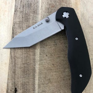 May include: A folding knife with a silver blade and a black handle. The blade is marked with the text "KA-BAR". The knife is open, displaying the blade's edge. The handle has a curved design for grip.