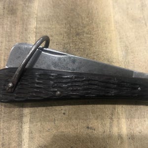 Pode incluir: Uma canivete vintage com cabo de madeira escura e lâmina de metal. A faca possui um laço de metal para prender a um chaveiro ou presilha. A lâmina está parcialmente aberta. A faca está sobre uma superfície de madeira.