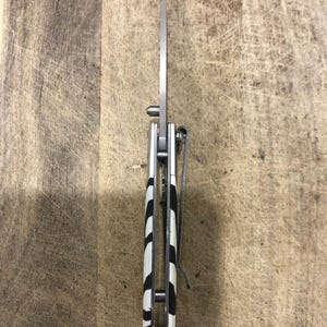 K&ouml;nnte beinhalten: Ein silbernes Klappmesser mit einer Klinge aus rostfreiem Stahl. Der Griff hat ein schwarz-wei&szlig;es Zebrastreifenmuster. Ein silberner Taschenclip ist am Griff befestigt.