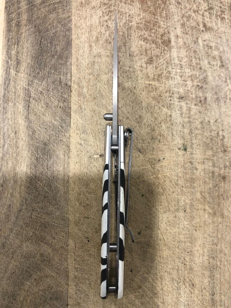 K&ouml;nnte beinhalten: Ein silbernes Klappmesser mit einer Klinge aus rostfreiem Stahl. Der Griff hat ein schwarz-wei&szlig;es Zebrastreifenmuster. Ein silberner Taschenclip ist am Griff befestigt.