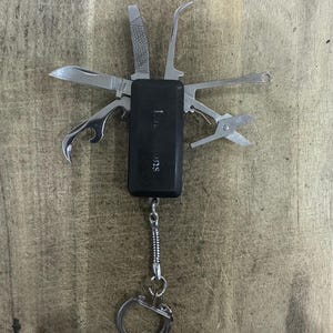 May include: A black multi-tool keyring featuring a rectangular handle with the text "SURVIVAL" printed on it. The tool includes several stainless steel implements extended outwards, such as a knife blade, a file, a hook, a bottle opener, and scissors.