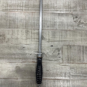 May include: A long, slender metal sharpening steel with a tapered, textured tip. The tool features a cylindrical black handle with horizontal ridges and a small metal guard separating the handle from the rod, plus a metal ring at the base.