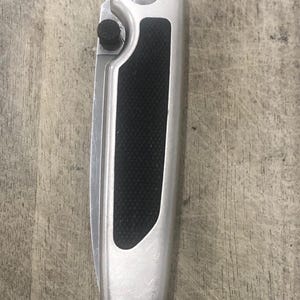 Pode incluir: Uma faca dobr&aacute;vel prateada com uma pega preta texturizada. A faca tem um bot&atilde;o na lateral e um elemento circular no topo. A l&acirc;mina est&aacute; fechada na imagem.